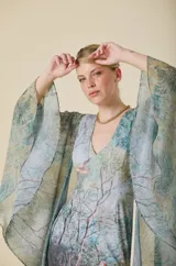 Vestido largo de corte sirena con escote en V, confeccionado en tela con estampado artístico de paisaje natural. Presenta mangas amplias y fluidas tipo capa en gasa semitransparente, que aportan movimiento y elegancia al diseño.