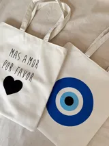 Tote bag de tela color blanco con estampado frontal de ojo turco en tonos azules y negros. Cuenta con doble asa de hombro, cierre principal y bolsillo interno.