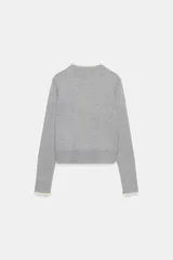 Chaqueta de punto de corte clásico, color gris, con cuello redondo y manga larga. Presenta cierre frontal con botones y detalles de puntilla en color crema en el cuello y los puños.