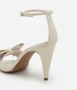 Sandalias de tacón alto color crema, con punta redonda abierta, tira fina con hebilla en el tobillo y maxi lazo decorativo en la parte delantera.