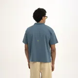 Remera tipo polo de manga corta, color verde oscuro, con cuello y abertura frontal. Presenta un estampado dorado en el pecho con el texto "PulauCrew" y un pequeño logo de corona de laurel bordado.