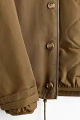 Campera de sarga levemente acolchada con cuello inglés, cierre frontal y solapa con botones. Presenta mangas raglán, bolsillos insertados delanteros y cordón ajustable en el bajo.