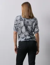 Blusa celeste con estampado blanco, de gasa, con cuello a la base y escote en V. Tiene abertura con botones, bolsillos delanteros y mangas 3/4 regulables con presilla y botón. El ruedo termina con un nudo.