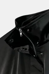 Cazadora satinada de color negro, con cuello subido, manga larga y bolsillos frontales de plastrón con cordones decorativos. Cuenta con bajo elástico y cierre frontal con cremallera y botones a presión oculta.
