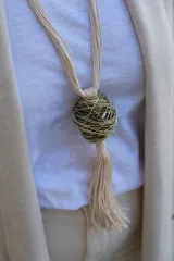 Collar largo con cordones de algodón color crema, que presenta un dije central esférico formado por hilos metálicos entrelazados en tonos dorados y plateados, finalizando con una borla de flecos de algodón.
