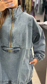 Buzo tipo canguro de denim con lavado medio, diseño oversize, cuello alto con cierre frontal, bolsillo delantero con cierre y bordado, puños elásticos y ruedo regulable.