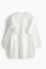 Vestido corto blanco de tejido liviano, con cuello redondo, botones forrados en el frente y mangas largas con puños finos abotonados. Presenta volados y aplicaciones de encaje en el canesú frontal.