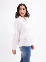 Camisa beige de manga larga con cuello camisero, cierre frontal con botones y diseño de pespuntes y bordados en relieve con formas florales en la parte inferior y lateral.