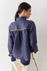 Camisa blanca de lino con cuello alto tipo gorguera, cierre frontal con botones de madera y detalles de ribetes en color mostaza en hombros y puños. Presenta volados en la botonera, cuello y puños.