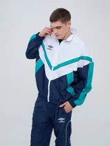 Campera deportiva Umbro modelo Beat, confeccionada en nylon ligero con diseño de bloques de color en blanco, azul marino y verde azulado. Presenta cierre frontal, cuello alto y elásticos en puños y cintura.
