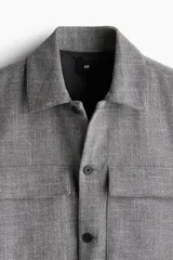 Sobrecamisa gris de corte recto, con cuello inglés, tapeta lisa y bolsillos superiores con solapa. Mangas largas con abertura y botón en los puños, canesú en la espalda y bajo recto.