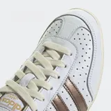 Championes Adidas modelo Japan confeccionados en cuero blanco con detalles metálicos en color bronce en las tres tiras laterales y el talón. Poseen un diseño de perfil bajo inspirado en los años 60, con puntera reforzada de doble costura, cierre de cordones y suela de caucho delgada.