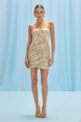 Vestido corto strapless dorado con textura metalizada. Presenta un corpiño ajustado con diseño floral en relieve y una falda drapeada cruzada que genera un efecto envolvente.