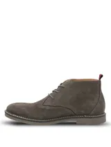 Botines tipo chukka Hush Puppies modelo New Safari, confeccionados en gamuza color gris. Presentan cierre de cordones, suela de goma con costura decorativa y detalle de tirador en el talón con franjas rojas y negras.