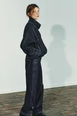 Cazadora de denim azul oscuro con cuello solapa, cierre frontal con cremallera y botones a presión ocultos. Presenta mangas largas tipo murciélago con puños, bolsillos de vivo en el delantero y bajo elástico ajustable con cordones.