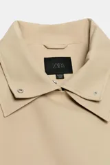 Trench corto oversize color beige con cuello subido de efecto doble, cierre cruzado con botones oscuros y bolsillos de vivo delanteros.