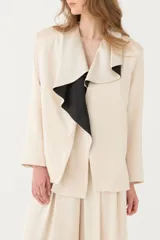 Blazer satinado color beige con hombros definidos, mangas largas y solapa con volado drapeado que cae sobre el frente, destacando un interior en contraste de color negro.