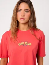 Remera coral cropped, de manga corta, con estampa trasera colorida de un perro surfeando y el texto "Rusty County Surf Club".