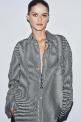 Camisa de corte fluido confeccionada en mezcla de viscosa, con diseño de rayas verticales en blanco y negro. Presenta cuello solapa, manga larga con puños, bolsillo de plastrón en el pecho, bajo redondeado y cierre frontal con botones.