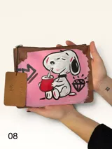 Sobre de cuero marrón con diseño pintado a mano que presenta al personaje Snoopy sobre un fondo rosado y la palabra 'heal'. Cuenta con cierre superior y etiqueta de cuero colgante.