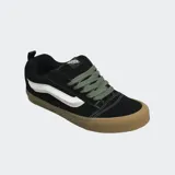 Championes Vans modelo Knu Skool, de color negro con la clásica franja lateral Sidestripe blanca. Presentan diseño robusto de estilo noventero, lengüeta y cuello acolchados, cordones anchos en color verde militar y suela de goma marrón.