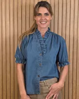 Camisa de denim de algodón celeste, con cierre frontal de botones, cuello alto con volados y mangas cortas abullonadas con puños elastizados fruncidos.