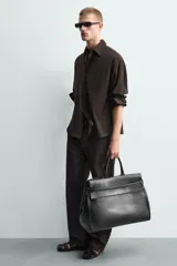 Bolso de mano extragrande negro de cuero sintético, con compartimento principal con cierre de imán, bolsillo interior con cierre y bolsillo secundario con solapa y detalle de cinta. Cuenta con dos asas de mano.
