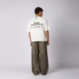 Remera boxy de color crudo con hombro caído y corte cuadrado a la cadera. En el frente presenta un diseño minimalista con texto y un pequeño logo central.