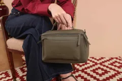 Bandolera de diseño rectangular en color suela, confeccionada en material sintético. Cuenta con un asa superior fija, correa larga ajustable y extraíble, y dos bolsillos frontales con cierre metálico, uno de ellos con diseño superpuesto. Presenta el logo de la marca grabado en el frente.