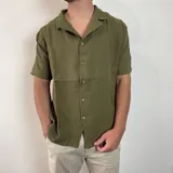 Camisa de hombre color verde oliva, de muselina con textura arrugada, manga corta, corte oversize, cuello cubano y cierre con botones. Presenta un pequeño logo bordado en el pecho.