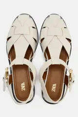 Sandalia plana tipo cangrejera color hueso, con tiras cruzadas y cierre mediante hebilla dorada.