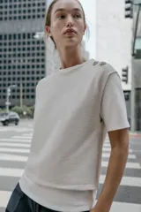 Top de cuello redondo y manga corta con diseño de capas que combina una remera de algodón blanca con un chaleco de punto gris superpuesto. Presenta detalles de botones joya en el hombro.