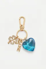 Llavero metálico dorado con gancho mosquetón y argolla, decorado con dijes colgantes: un trébol de cuatro hojas con pedrería, un pequeño animal alado, una placa con la palabra 'LOVE' y un gran corazón azul translúcido de acrílico.