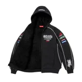Campera Supreme de estilo racing en color negro, con capucha forrada en corderito negro, cierre frontal completo y bolsillos tipo canguro. Presenta diversos parches bordados con temática automovilística en el pecho y las mangas, además de líneas blancas decorativas.