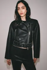 Chaqueta tipo biker de piel negra con solapas, cierre frontal asimétrico con cremallera, bolsillos con cierre y cinturón ajustable con hebilla metálica en la cintura.