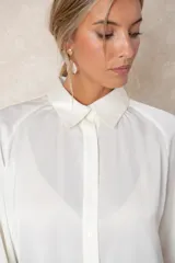 Blusa de satén color marfil con brillo sutil, cuello clásico, cierre frontal con botones y mangas largas con puños abotonados. Presenta un corte fluido y elegante.