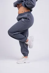 Pantalón de jogging gris oscuro, de corte holgado con cintura elástica y puños ajustados en los tobillos. Presenta bolsillos laterales y un pequeño logo bordado en la pierna izquierda.