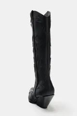Bota estilo cowboy de caña alta, color negro, con detalles en relieve y costuras decorativas. Presenta punta fina, tiradores laterales y taco cuadrado inclinado.