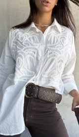 Camisa blanca de corte oversize con diseño de bordado calado en el frente. Presenta cuello clásico, cierre frontal con botones y mangas largas.