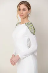 Vestido largo blanco de manga larga, con escote redondo al frente, espalda baja descubierta y detalles de bordado de piedras y pedrería en tonos verdes sobre los hombros.