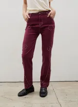 Pantalón de denim color bordó, de corte recto y ajuste cómodo, decorado con tachas metálicas dispuestas en patrones circulares y líneas laterales. Presenta costuras en contraste y cierre frontal con botón.