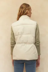 Chaleco puffer color crema, con cuello alto, cierre frontal y cuatro bolsillos con solapa. Cuenta con cintura ajustable con cordón.