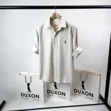 Camisa de vestir color beige claro, de cuello italiano y manga larga. Tiene un pequeño logo bordado en el pecho.