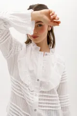 Camisa blanca de algodón con diseño romántico, cuello alto con volados, cierre frontal con botones nacarados y mangas largas con puños acampanados. Presenta detalles de encaje y franjas horizontales en toda la prenda.