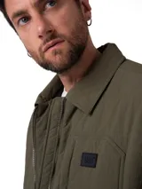 Chaqueta de hombre color verde oliva, con cierre frontal de cremallera, cuello camisero, bolsillos frontales tipo parche y parche de logo en el pecho. Presenta un diseño funcional con tecnología resistente al agua y al viento.