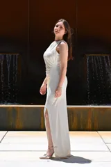 Vestido largo de fiesta color gris claro metalizado, con escote redondo, sin mangas y abertura lateral en la falda. Presenta un drapeado lateral que estiliza la figura.