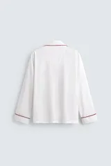 Pijama de dos piezas confeccionado en popelín de algodón blanco, con vivos de ribete en color bordó. Incluye una camisa de manga larga con cuello bowling, bolsillos de plastrón y cierre frontal de botones, y un pantalón de corte relajado con cintura elástica y bolsillos laterales.