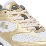 Championes Lacoste modelo Storm 96 2K, con diseño deportivo de estilo retro. Presenta una combinación de malla blanca y paneles superpuestos en tono dorado metalizado, con el logo del cocodrilo en el lateral. Suela robusta con detalles en gris y negro.