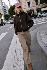 Chaqueta estilo bomber de corte oversize en color marrón oscuro, confeccionada en tejido suave. Presenta capucha con cordones ajustables, cierre frontal con cremallera, mangas largas con puños elásticos y bolsillos delanteros de vivo. El bajo cuenta con detalle de pliegues.