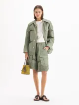 Chaqueta de gabardina liviana color verde militar, con silueta recta, largo por debajo de la cadera y cuello de corderoy. Presenta detalles de encaje en hombros, mangas y ruedo, múltiples bolsillos con solapa y cierre frontal con broches metálicos.